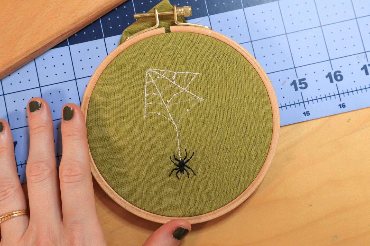 an embroidery of a spider dangling from it's web stitched on green fabric, framed in an embroidery hoop