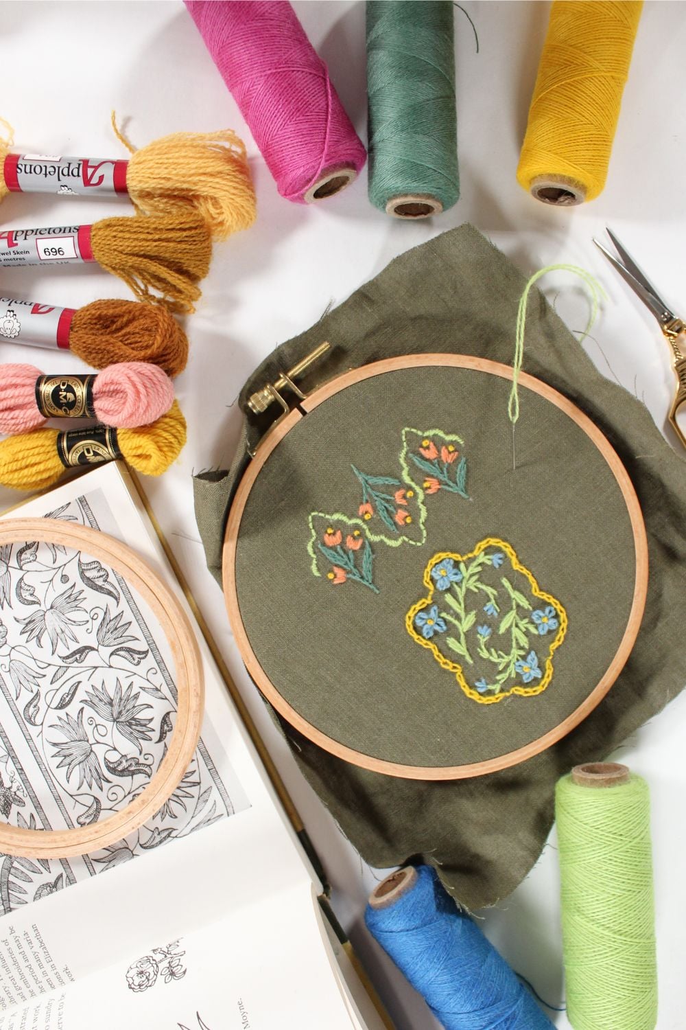 crewel embroidery on dark green linen fabric surrounded by embroidery supplies.