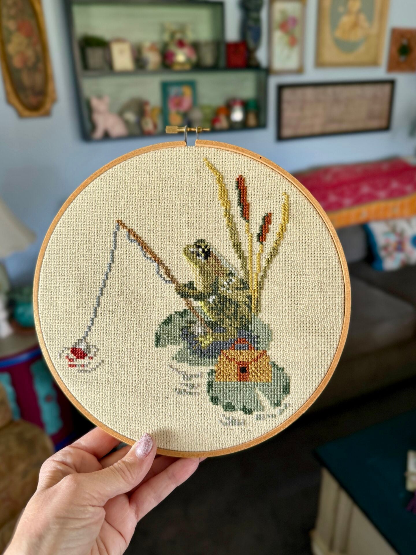 a hand holding a cross stitch design of a frog sitting on a lily pad, holding a fishing pole. Beside him is a tackle box and behind the frog are some cattails.