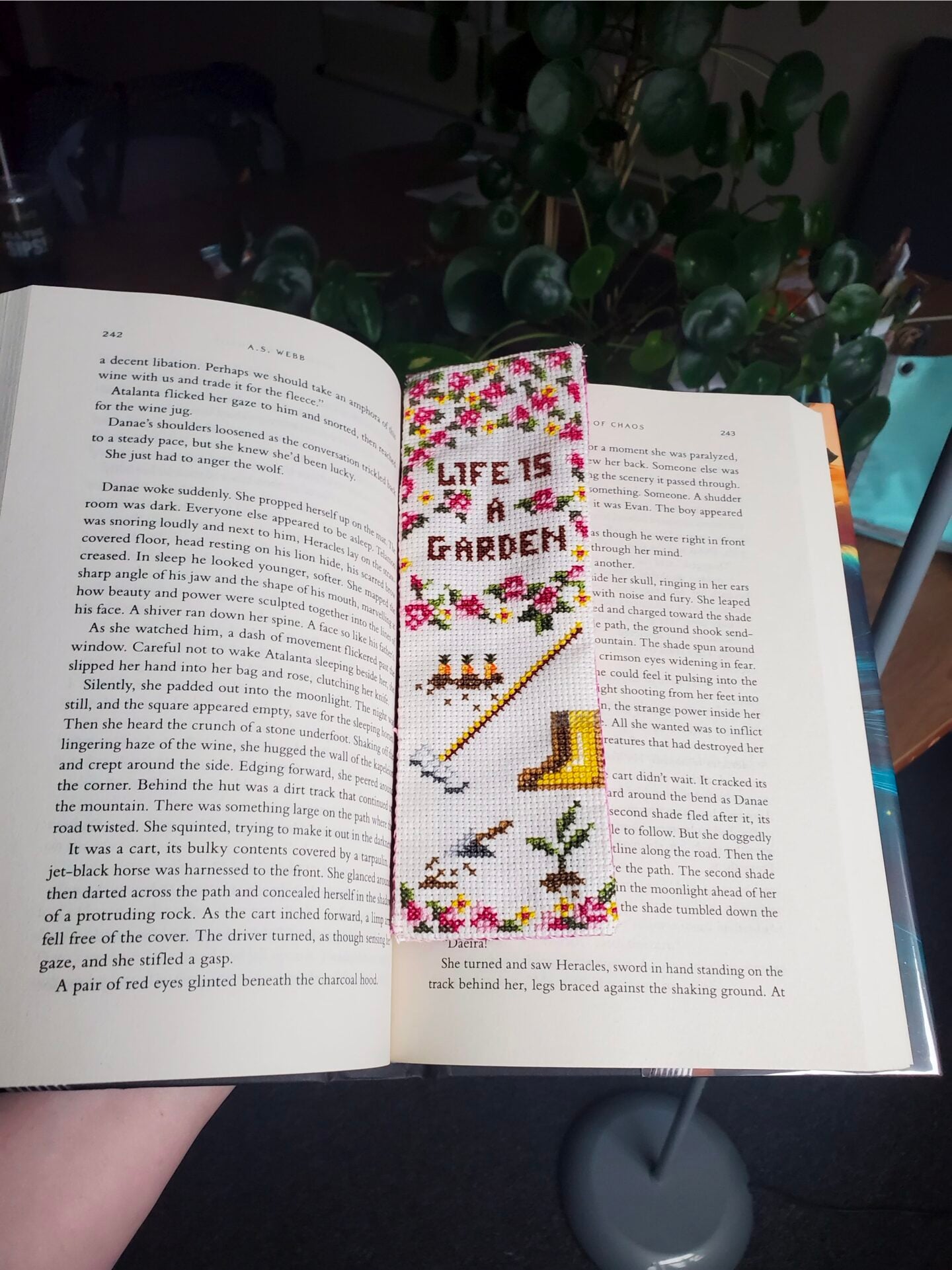 cross stitch bookmark laying in an open book. The cross stitch design features the quote "life is a garden" with pink and yellow flowers, carrots in the ground, and some gardening tools