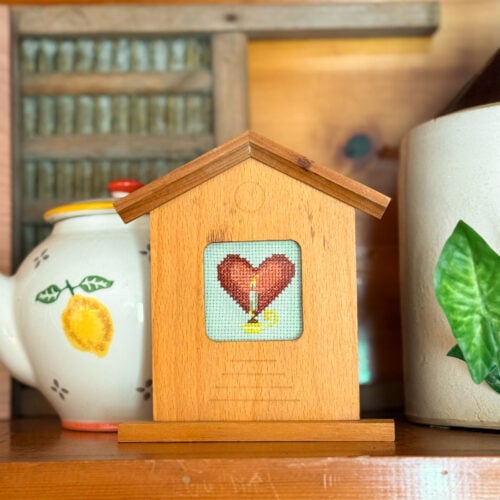 cross stitch of a burning candle and red heart. The embroidery is framed in a wood frame shaped like a house that is sitting on a shelf near a teapot and washboard in the background.