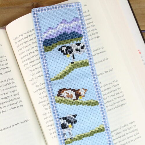 a cross stitch bookmark of black and brown cows with mountains in the background. The bookmark has a gingham border and is stitched on light blue Aida cloth.