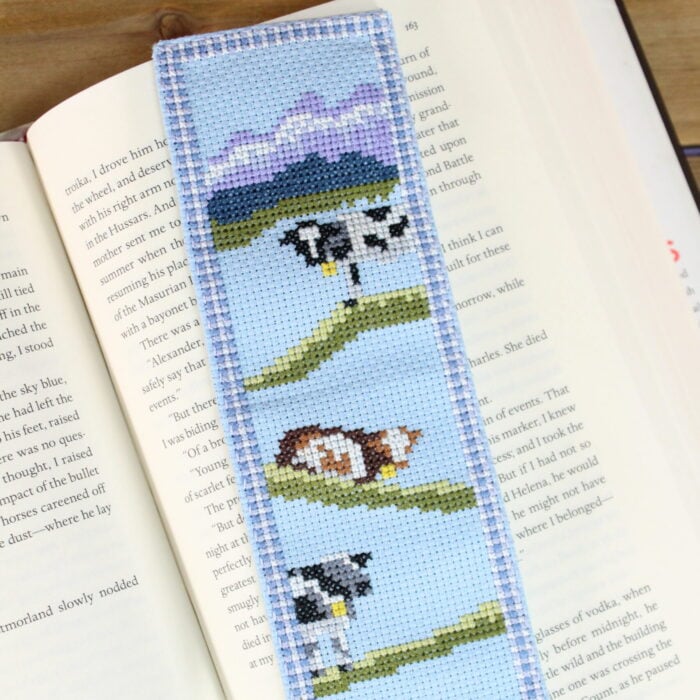 a cross stitch bookmark of black and brown cows with mountains in the background. The bookmark has a gingham border and is stitched on light blue Aida cloth.