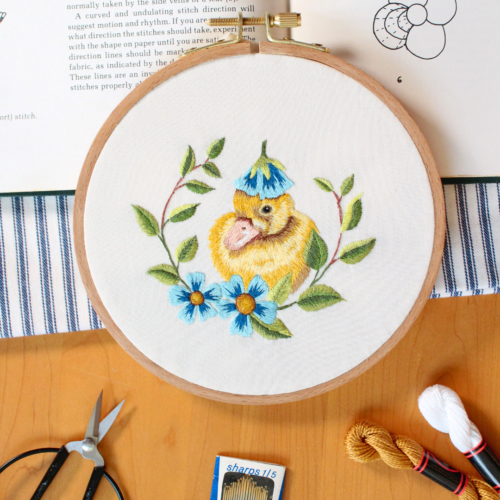 embroidery of a yellow duckling with a blue flower on it's head and blue flowers and leaves surrounding it