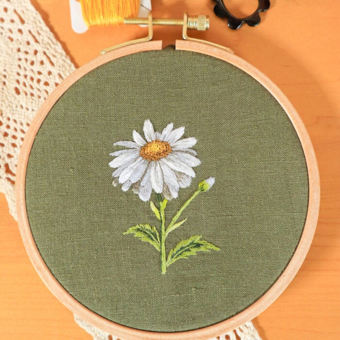 an embroidery of a daisy flower stitched on dark green linen fabric. The embroidery is framed in a light wood embroidery hoop and laying on a wood surface