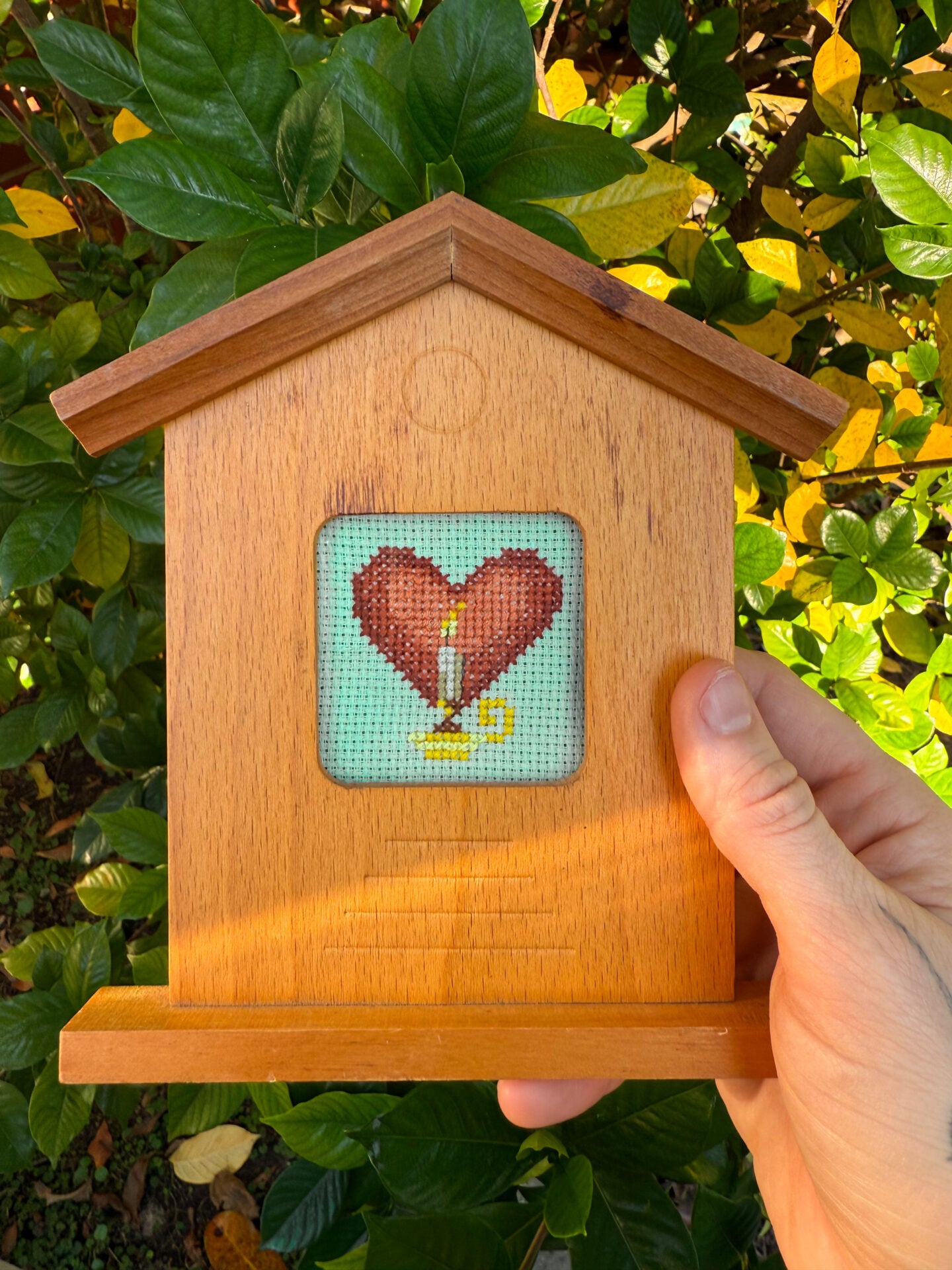 small heart and candle cross stitch pattern framed in a wood frame shaped like a birdhouse