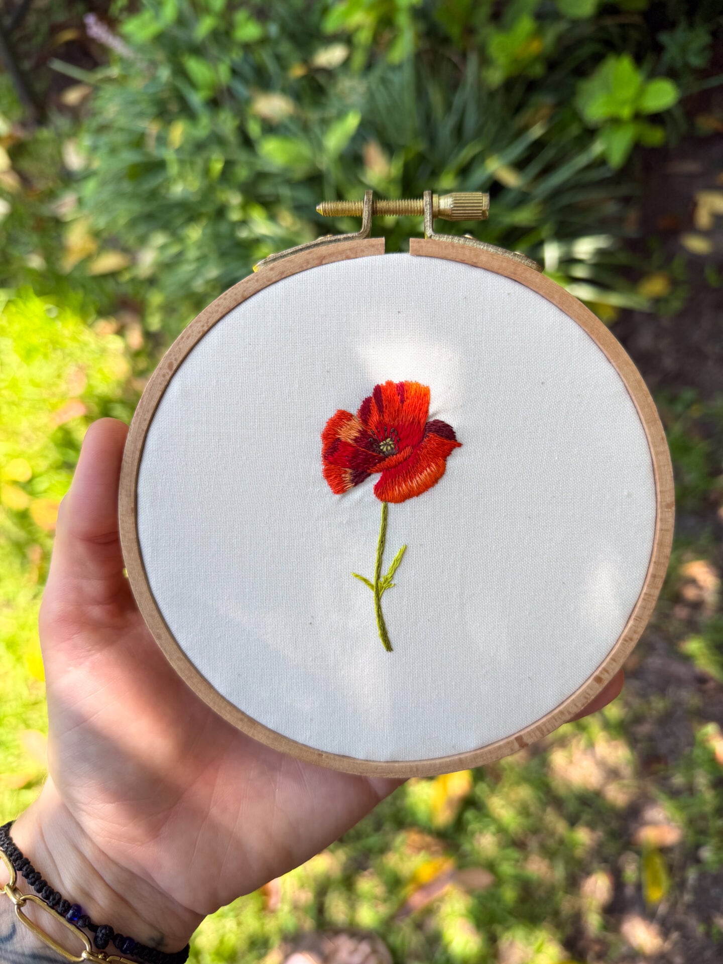 embroidery of a red poppy flower on off white fabric, framed in a wooden embroidery hoop