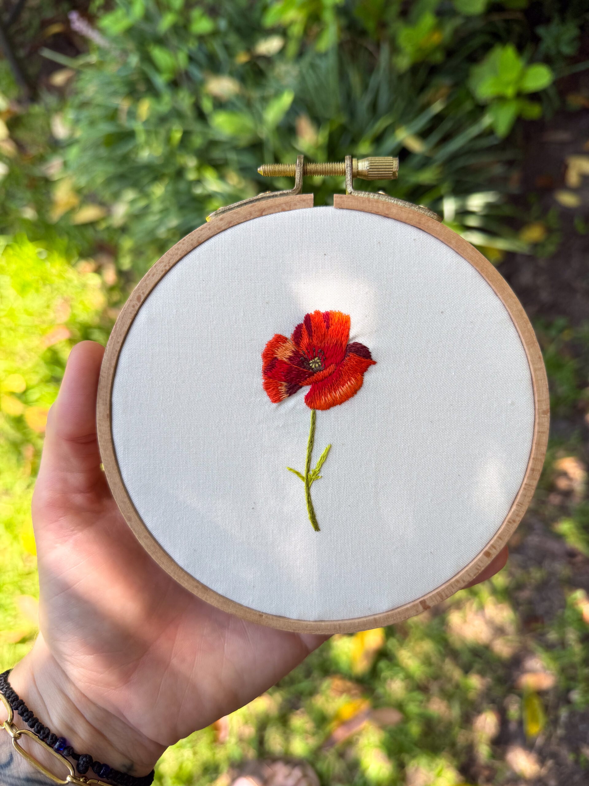 embroidery of a red poppy flower on off white fabric, framed in a wooden embroidery hoop